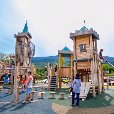A KOMPAN wooden castle-themed play structure with slides and climbing areas, surrounded by children and families playing.