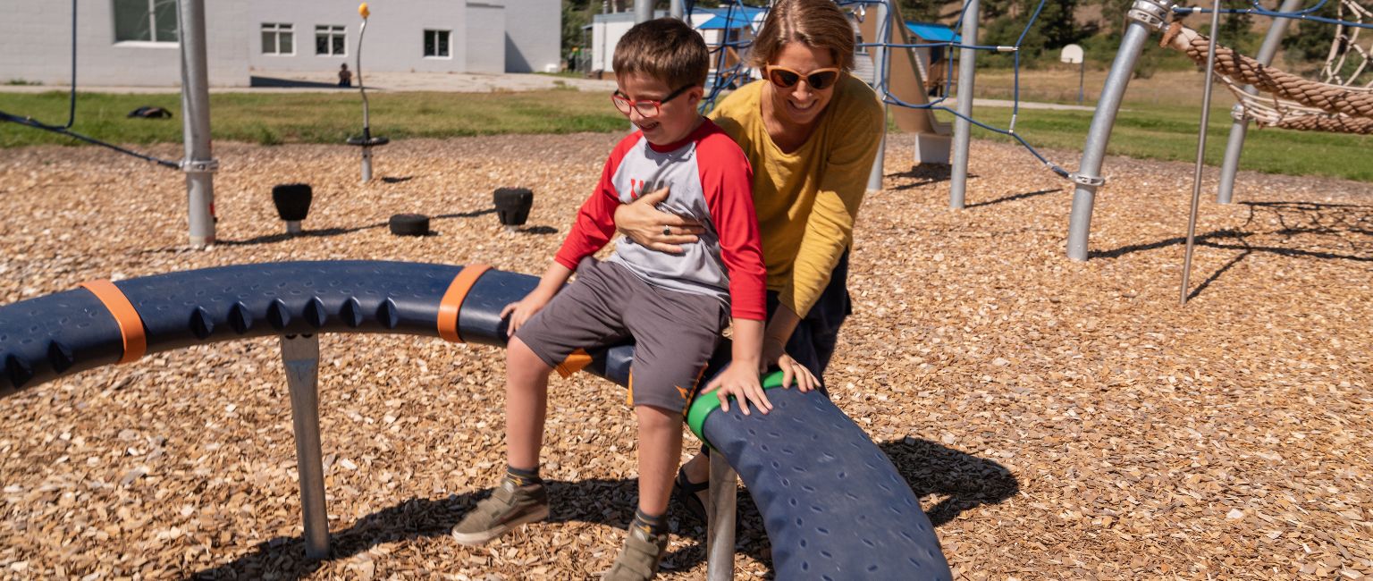 2 Young boy and caregiver playing on a KOMPAN playground spinner on engineered wood fibre surfacing
