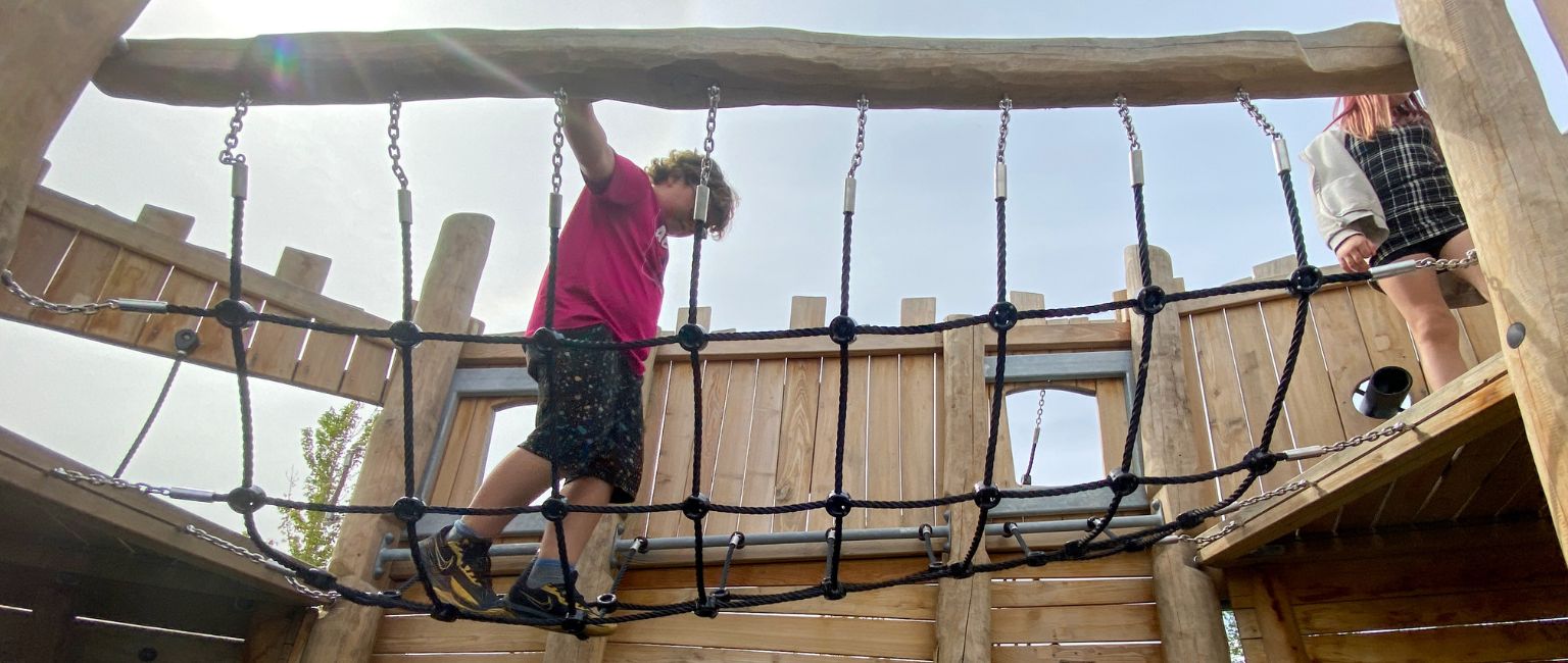 A young boy uses balance and coordination to walk across a rope ladder that is part of a larger playground structure