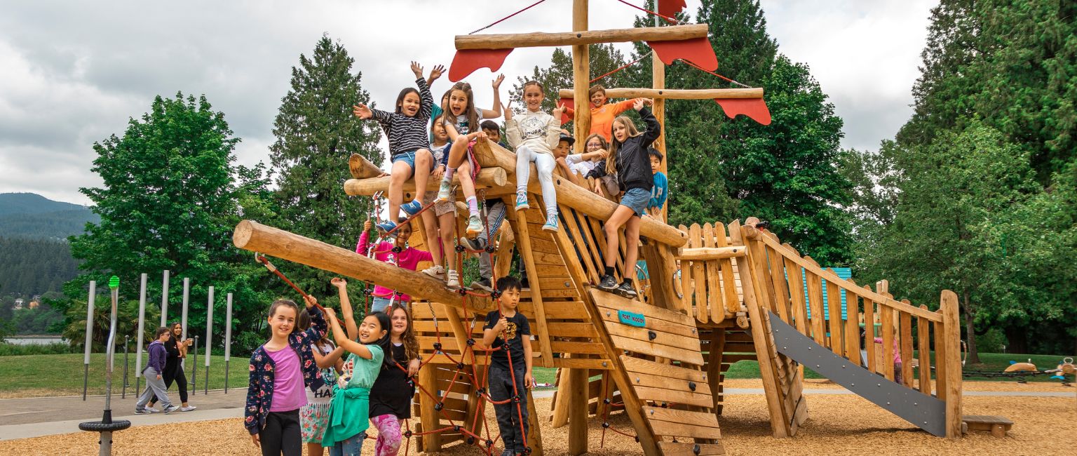 A group of kids play on a massive KOMPAN Robinia pirate ship structure on the playground.