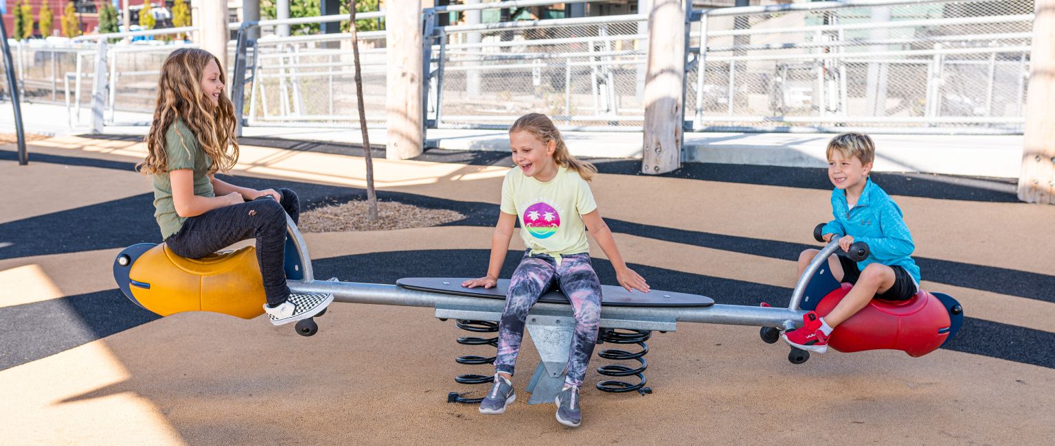 Three kids playing on a seesaw at a playground while laughing
