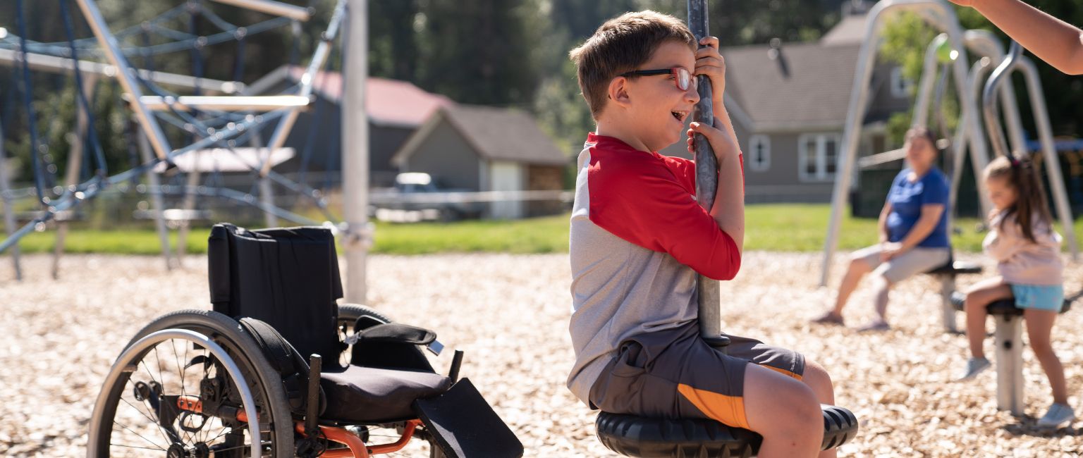 Young boy plays on an inclusive and accessible playground spinner with his wheelchair in the background